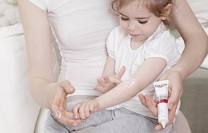 Djevojčica sjedi u majčinu krilu, nanosi Eucerin AtopiControl Akut kremu na pregib svoje ruke.